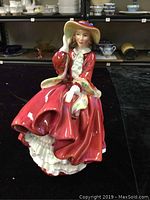 Front view of the Royal Doulton Top of the Hill figurine showing the woman in a red dress holding her wide-brimmed hat.