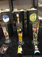 Three Ritzenhoff pilsner glasses shown upright in front of their original black cylindrical packaging tubes, each tube and glass featuring a unique design corresponding to one of the glasses.