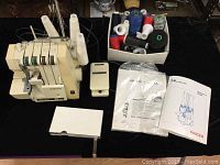 Overall lot including serger machine, foot pedal, manuals and box of thread spools on black surface