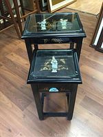 Two black lacquered nesting tables, the larger one on top, showing inset glass tops with hand-painted stone figurines and gold painted landscape designs.