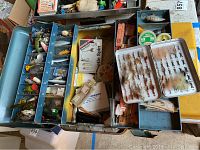 Overview of two metal tackle boxes filled with fishing lures and related items including packaged lures, fishing line spools, and other tackle items.