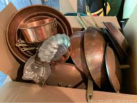 Box containing various copper pots, pans and molds with metal handles and some patina/oxidation.