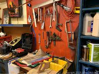 Photo shows a workspace with assorted hand tools including wrenches, saws, pliers, screwdrivers, an extension cord hung on a red pegboard, work light, and other workshop supplies.