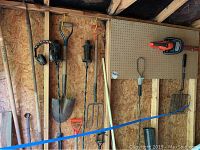 Photo showing a variety of yard tools including shovels, pitchfork, garden fork, and ear protector headphones hanging on wall and leaning on wall.