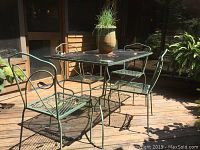 Green wrought iron patio table with rectangular glass top surrounded by four matching chairs on outdoor wooden deck under sunlight.