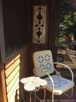 Two vintage metal outdoor chairs with cream-colored metal seats and backs featuring black decorative cutouts, and matching metal side table with butterfly-shaped tile top, situated on a wooden deck.