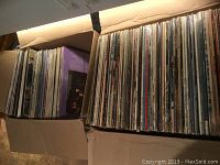 Two cardboard boxes filled with a large assortment of vinyl LP records packed upright side by side showing spines.