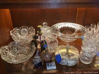 Assortment of perfume bottles, various cut glass dishes and servingware including bowls, decanters, and small glasses displayed on wooden shelving.
