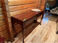 Side angle view of the console table against a log cabin style wooden wall, showing Queen Anne style legs and polished wood surface.
