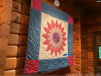 Handmade quilt hanging on a wall showcasing a starburst pattern in red, cream, and blue with quilted designs on the borders