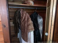 View of three fur coats hanging in a wooden closet including a grey muskrat coat, white mink coat, and a black garment cover