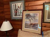 Two prints displayed, one depicting redhead ducks among trees with wood log wall background, the other of ducks flying over water and reeds, showing framing and matting.