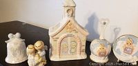 Five ceramic Precious Moments figurines displayed on table, including church-shaped keepsake, bell, decorative plate, and two additional figurines