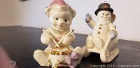 Photo showing Lenox 1999 teddy bear playing a drum and snowman in sleigh figurines side by side.