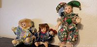 Photo of three porcelain dolls set including jester doll, clown doll, and small girl doll, displayed against a wall and on surface.