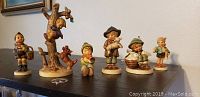 Wide shot showing all six Hummel boy figurines lined up on a shelf with a white wall background.