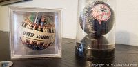 Photo showing two baseballs in original clear plastic packaging, one square case with stadium design and one round case with Yankees logo