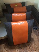 Four square pillows with an orange leather strip center and dark fabric sides arranged on a hardwood floor and leaning against a wall.