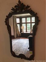 Full view of ornate wall mirror hanging on wall reflecting room interior with window and furniture.