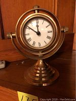 Front view of brass ship's clock with white face, Roman numerals, black hands, mounted in a globe-like brass frame with adjustable knobs, on a stepped brass base.