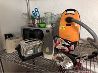 Children's toy appliances including two toasters, microwave, blender, iron, vacuum cleaner, kitchen utensils, mixer whisks and fabric oven mitt arranged on metal shelving.