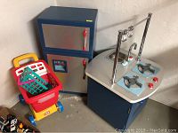 Wide shot showing Pottery Barn Kids play refrigerator/freezer unit next to a play shopping cart with green baskets and toy food items inside.
