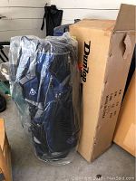 Photo showing Dunlop golf bag still wrapped in clear plastic standing next to its box labeled with Dunlop logo, indicating the bag is new and unused.