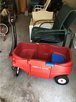 Laser shipper red plastic Radio Flyer wagon with blue seat and black handle seen from side with wheels and door visible in garage setting