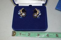Pair of blue and clear rhinestone clip on earrings in blue velvet box with measuring tape showing size.