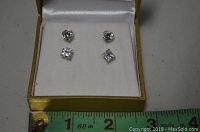 Two pairs of stud earrings set in a white cushioned box with yellow trim, showing round and square clear stones.