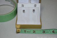 Two pairs of stud earrings in a box with a green tape roll and measuring tape for scale, showing approximate size of earrings