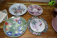 Four collector plates with floral and bird designs on a wooden table, some including certificates.