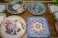 Set of seven collector plates arranged on wood surface showing front designs, some with accompanying certificates of authenticity.