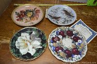 Photo showing four collector plates on wooden surface: white rose, birds on branch, red apples with flowers, floral bouquet with roses.