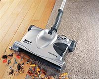 Cordless sweeper in silver and black showing the brush head on a hardwood floor with visible debris.