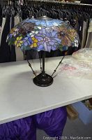 Full view of stained glass Tiffany-style lamp showing colorful floral glass shade and dark metal base on table in front of hanging clothes.