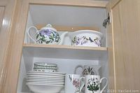 Overall view of cabinet shelf displaying teapot and covered casserole on top shelf with mugs, pitcher and bowls below