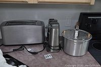 Three stainless steel kitchen appliances including toaster and can opener with a Whirley Pop popcorn maker to the right on countertop.