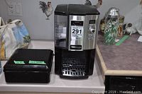 Photo showing Cuisinart coffee maker next to a black box on a kitchen counter with some background items.