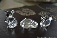 Three Swarovski crystal figurines displayed on a dark surface: a koala bear on the left, a clam shell in the center, and a swan on the right.