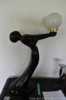 Black tall figurine lamp shaped as an elongated female figure in Art Deco style holding a white globe lamp shade on a black base.