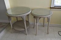 Two beige wicker tables with round glass tops, one larger with lower shelf and one smaller without shelf, showing the overall look and condition.