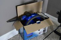 Blue handheld steam cleaner with attached hose inside an open cardboard box on carpeted floor.