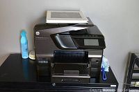 Front angled view of the black HP Officejet Pro 8600 Premium printer on a black cabinet, with a spray bottle on the left and a white binder on top.
