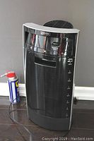 Black Bionaire humidifier standing on dark floor next to a can of WD-40, view of front and side showing size and shape.