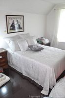 Double bed in a bedroom with white and light gray patterned bedding, multiple pillows, and wooden nightstand visible.