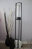 Tall black metal candle holder with glass shade, black and white decorative sticks in black clay vase, flameless candles on the floor.
