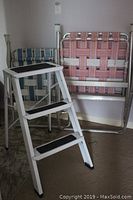 Pair of aluminum framed lawn chairs with woven seats and backrests in blue and pink colors, displayed with a white metal 3-step stool with black treads.