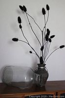 Photo of the five gallon glass vessel lying horizontally and the clay vase with teasel stems placed on a wooden surface against a white wall.
