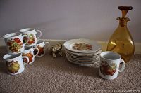 Full set of nine dessert plates stacked, five coffee mugs stacked and arranged, gold-toned elephant figurine, and amber glass decanter with stopper on carpet.